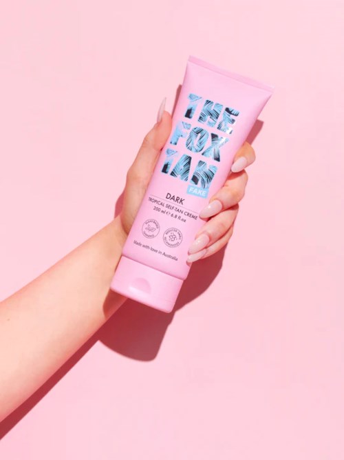 En hånd som holder en rosa tube med hudkrem mot en rosa bakgrunn.