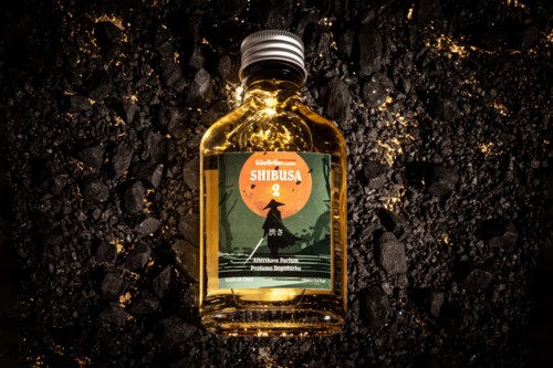 Kleine bernsteinfarbene Parfümflasche mit einer Illustration eines Mädchens auf einem Hügel bei Sonnenuntergang, auf dunklem, strukturiertem Boden.