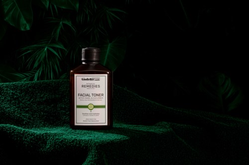 A dark brown bottle labeled 'Facial Toner' sits on a textured green surface with tropical plants in the background.