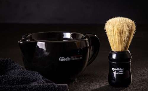 En svart rakskål och borste med en svart handduk, alla med Goodfellas smile-logotypen, på en mörk yta.