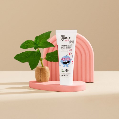 En børnetandpasta tube med jordbærsmag ved siden af en lille grøn plante i potte, med en pink dekorativ bue bagved.