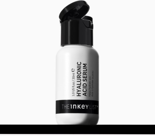 En hvit flaske med svart lokk, merket Hyaluronic Acid Serum av The Inkey List, står på en mørk overflate.