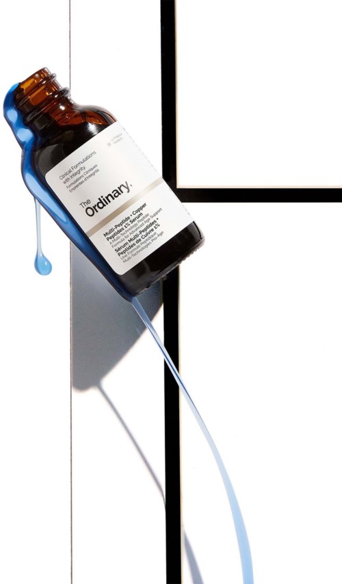 A brown glass bottle labeled 'The Ordinary' tilted on a white door, with a blue drop of liquid falling from it.
