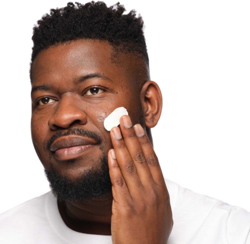 A man applies skincare cream to his cheek, smiling slightly. He has dark, curly hair and facial hair.