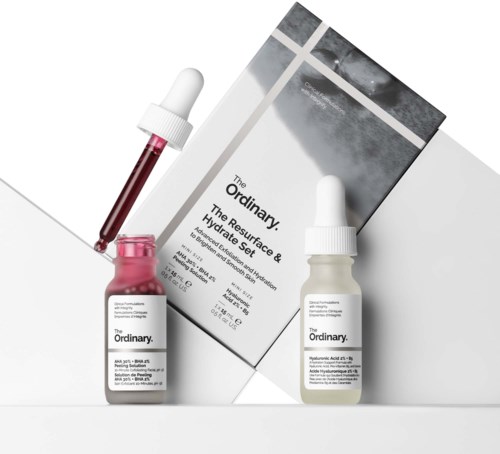 Two bottles of skincare serum with dropper caps are placed in front of a box, which displays the brand's name and product info.