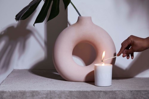 Someone lighting a candle near a white decorative vase with a large hole, on a gray surface with a plant in the background.