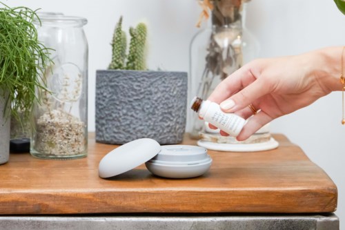 A hand drops liquid into a container on a wooden table with plants and jars nearby.
