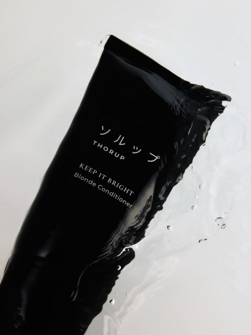 Schwarze Tube mit Blonde Conditioner und japanischem Markennamen, liegt auf weißer Oberfläche mit Wasser.