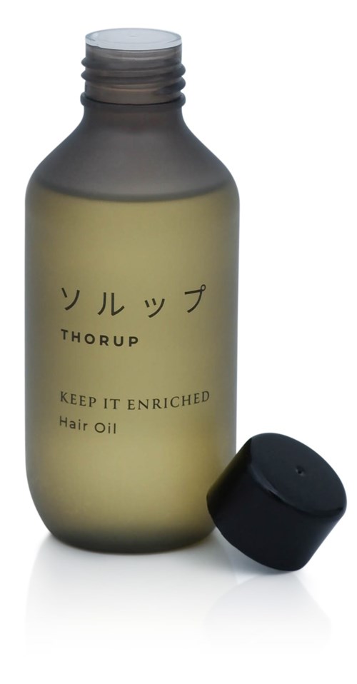 En grå flaska märkt 'Thurup Keep It Enriched Hair Oil' med en svart lock bredvid.