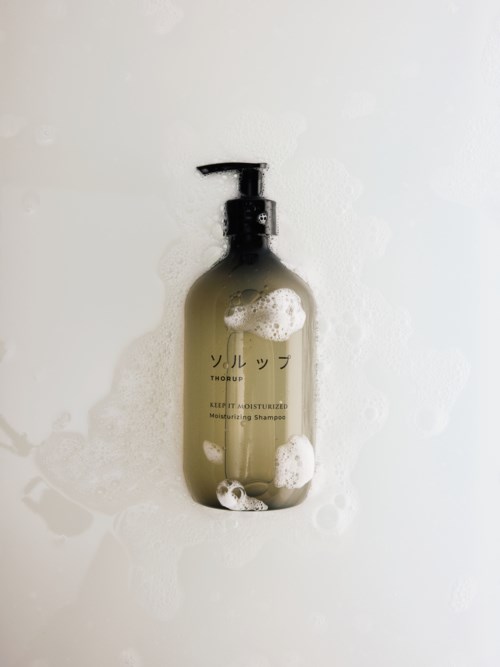 Eine beige Flasche mit schwarzem Pumpkopf, auf Japanisch beschriftet, im Duschbereich mit Seifenschaum.