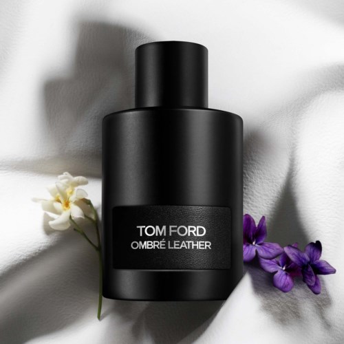 En svart flaska av Tom Ford Ombré Leather-parfym på vitt tyg, omgiven av lila blommor.