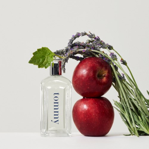 A bottle labeled 'Tommy' next to two red apples and a bunch of purple flowers with green leaves.