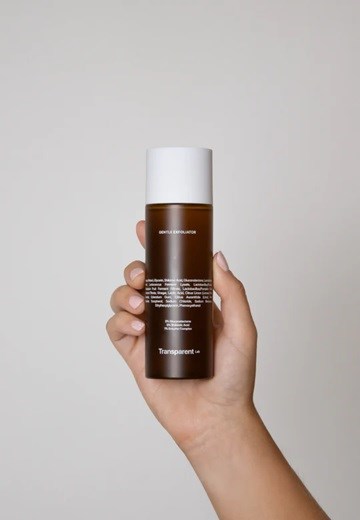 A hand holds a brown skincare bottle with white cap, with text on the label and a plain gray background.