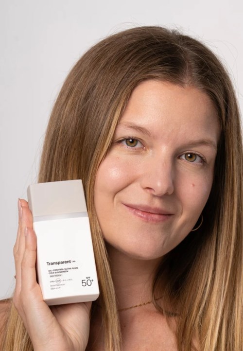 A woman with long hair smiling and holding a white package with text, showing it to the camera.