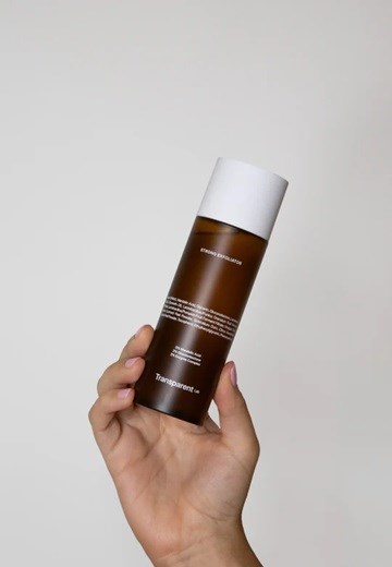 Eine Hand hält eine braune und weiße zylindrische Flasche vor neutralem Hintergrund.