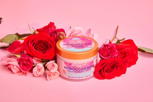 Een potje Moroccan rose whipped shea body butter omgeven door roze en rode rozen op een roze achtergrond.