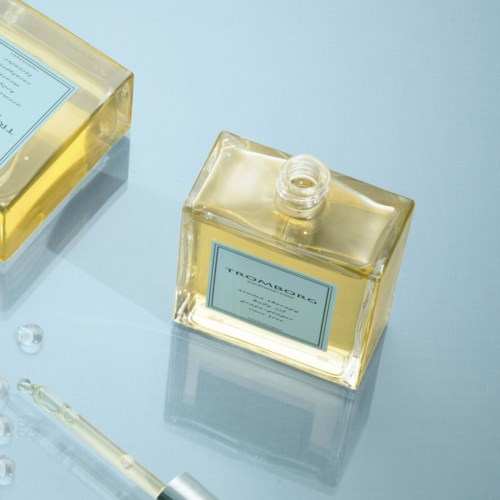 En klar, rektangulær parfume flaske med label, liggende på en lyseblå overflade, ved siden af en mindre flaske og pipette.