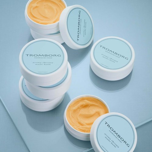 Otte beholder med gulagtig salve eller creme, mærket 'TROMBORG' med blå låg, arrangeret på en blå overflade.