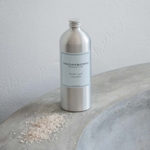 En sølvlignende flaske mærket 'Tromborg bath salt vanilla' står på en grå overflade med lidt salt spredt i nærheden.