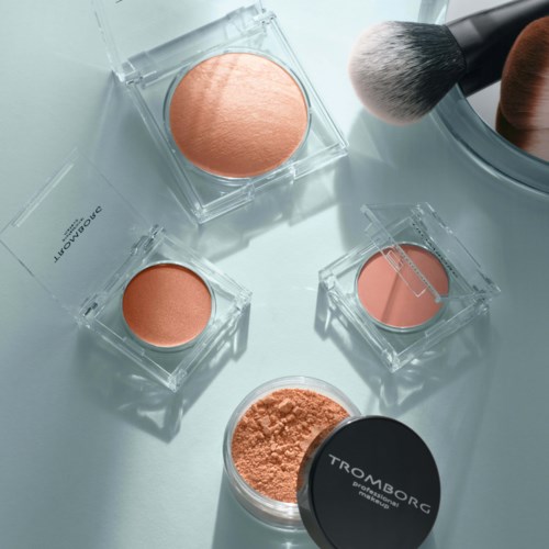 En samling makeup produkter, herunder pudder, blush og børster, arrangeret på en hvid overflade.