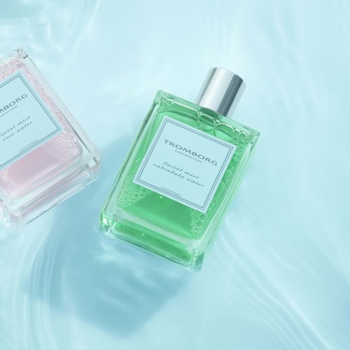 To parfume-flasker, en grøn og en pink, ligger på en blank overflade. Den grønne flaske er tydelig og i forgrunden.