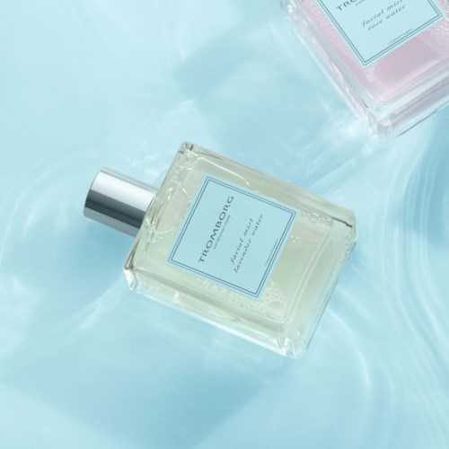 To parfume flakoner ligger på en lys blå overflade og reflekterer forsigtigt. Den ene er klar, den anden er pink. Begge har sprayhætter.