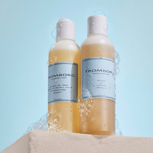 To shampoo-flasker med blå mærker står på en overflade, omgivet af sæbe bobler, mod en lyseblå baggrund.