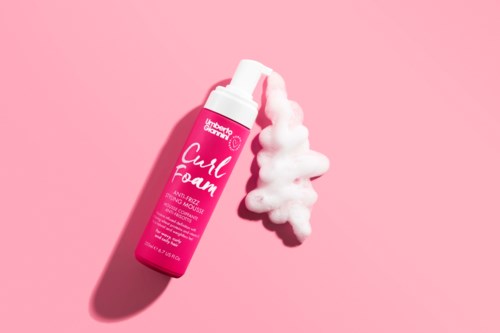 En rosa flaska med labeln 'Cush Foam' med skum som rinner ut på en rosa bakgrund.
