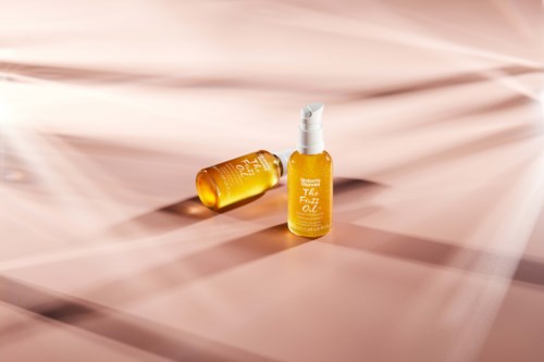 Två gyllene flaskor med spraylock är på en mjuk rosa tygbakgrund.
