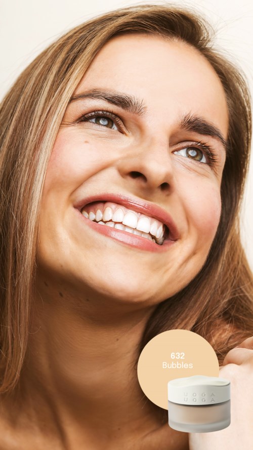 En kvinde med langt blondt hår smiler glædeligt, viser sine tænder, mod en enkel baggrund.