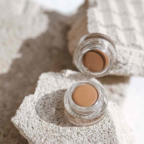 Two small jars of beige cream placed on textured, white stones, with a towel in background.