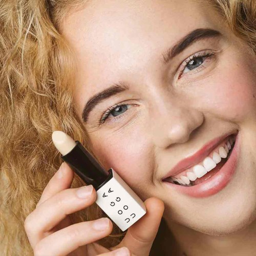 A young woman with curly hair smiling and holding a lipstick close to her face.