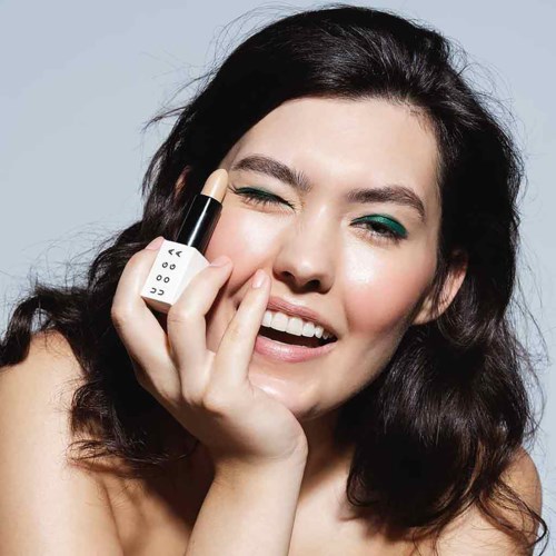 A woman with dark hair applying makeup, smiling, and holding a lipstick close to her face.