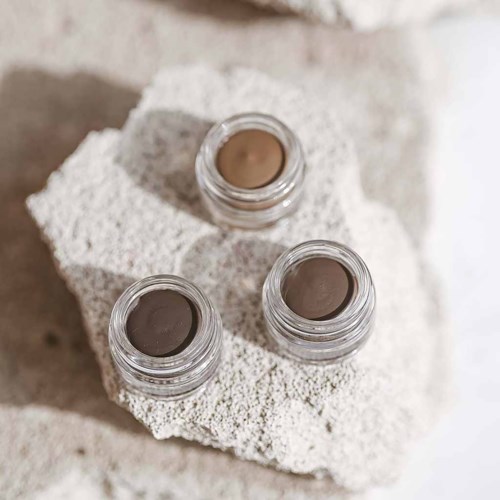 Tre små krukker med makeup eller gelé på en hvid tekstureret overflade med neutral baggrund.