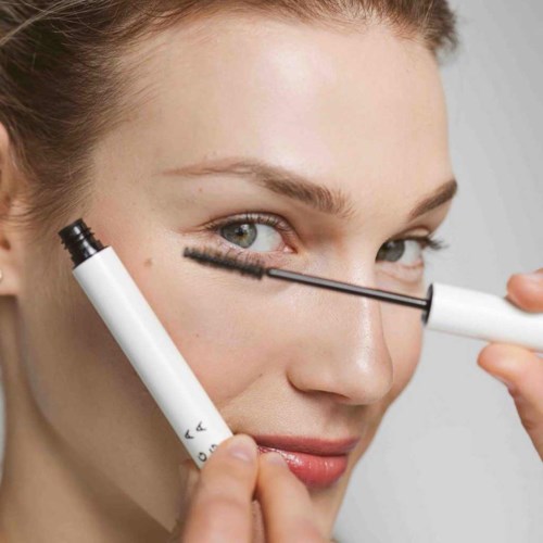 A woman applies mascara to her eyelashes, holding a mascara tube and brush.