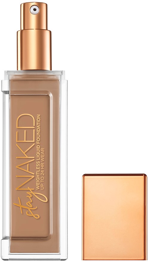 En foundationflaske med spraydeksel og gullhatt, merket "Stay Naked," i en gjennomsiktig beholder.