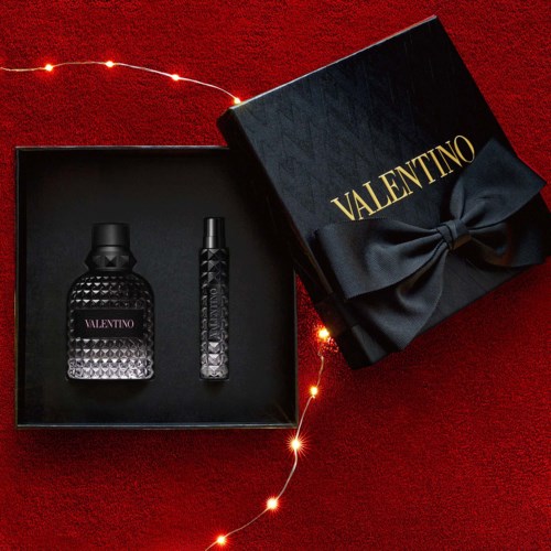 A Valentino perfume gift set in a black box with gold logo and a ribbon, on a red surface with fairy lights.