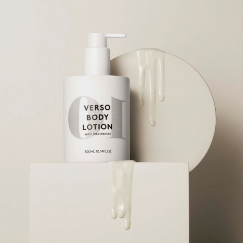 En vit flaska med kroppslotion med pump, märkt "Verso Body Lotion," på en vit yta med lotiondroppar.