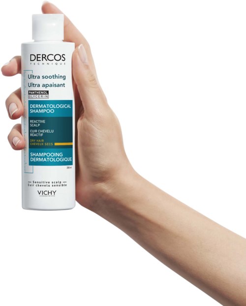 A hand holds a white bottle of Dercos shampoo, which is designed for sensitive scalp and dry hair.