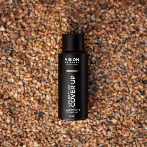 En svart burk märkt 'VISION HAIRCARE' och 'COVER UP' placerad på små, färgglada stenar.