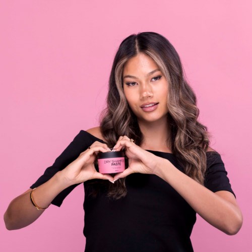 En kvinde med langt, bølget hår holder en pink krukke mærket "dry shampoo paste" mod en pink baggrund.