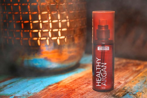 En röd sprayflaska märkt 'Healthy Argan' på en träyta, med en stor metallkittel i bakgrunden och ånga eller rök som stiger.