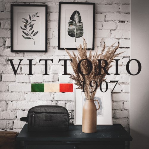 Inredning med inramade lövbilder, ett tecken med 'VITTORIO', Italiens flagga och en vas med torkade växter på ett svart bord.