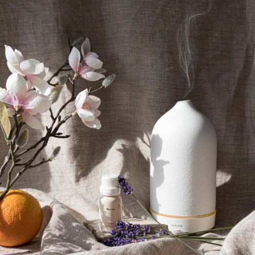 A white diffuser emits vapor, with pink and white flowers, an orange, and purple lavender resting nearby on a beige fabric.