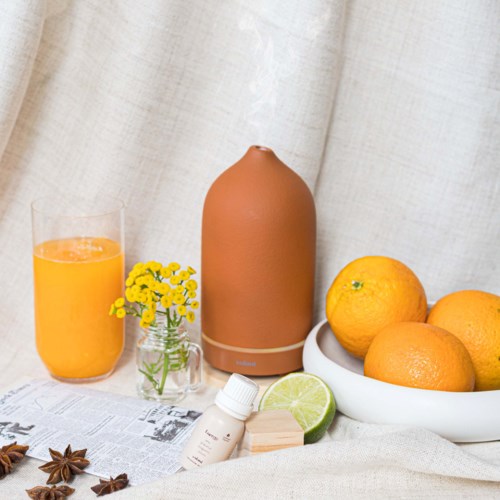 A orange juice glass, a small yellow flower vase, a tan humidifier, oranges in a white bowl, and a lime half.