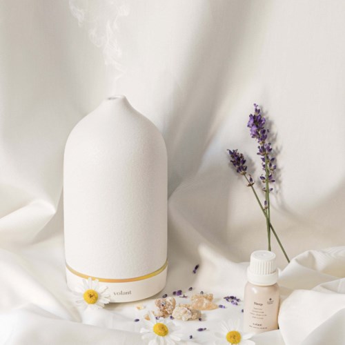 A white, rounded diffuser with a small yellow flower, a tiny bottle, and lavender sprigs on a white fabric background.