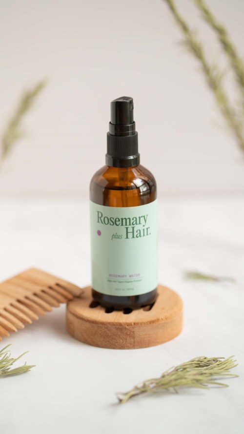 Bernsteinfarbener Sprühflasche mit 'Rosemary plus Hair'-Etikett. Mit Holzstück und Rosmarinzweigen.