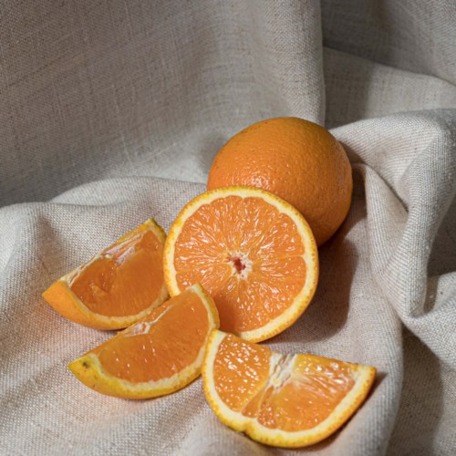 Orange fruit, whole and cut, on beige fabric, showing juicy inside and textured skin.