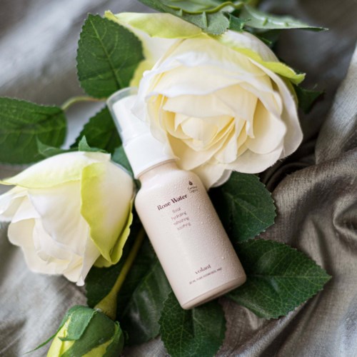 A bottle of rose water lies among white roses and green leaves on a soft surface.