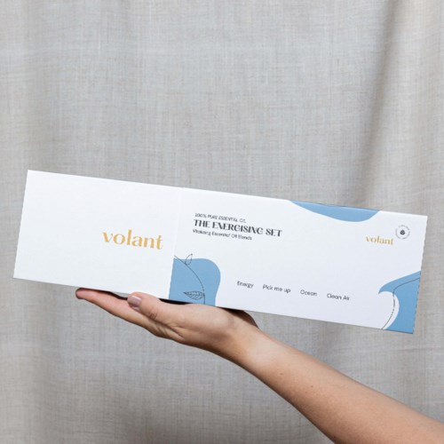 A hand holds a long, white box with blue and orange text, lying against a beige curtain background.
